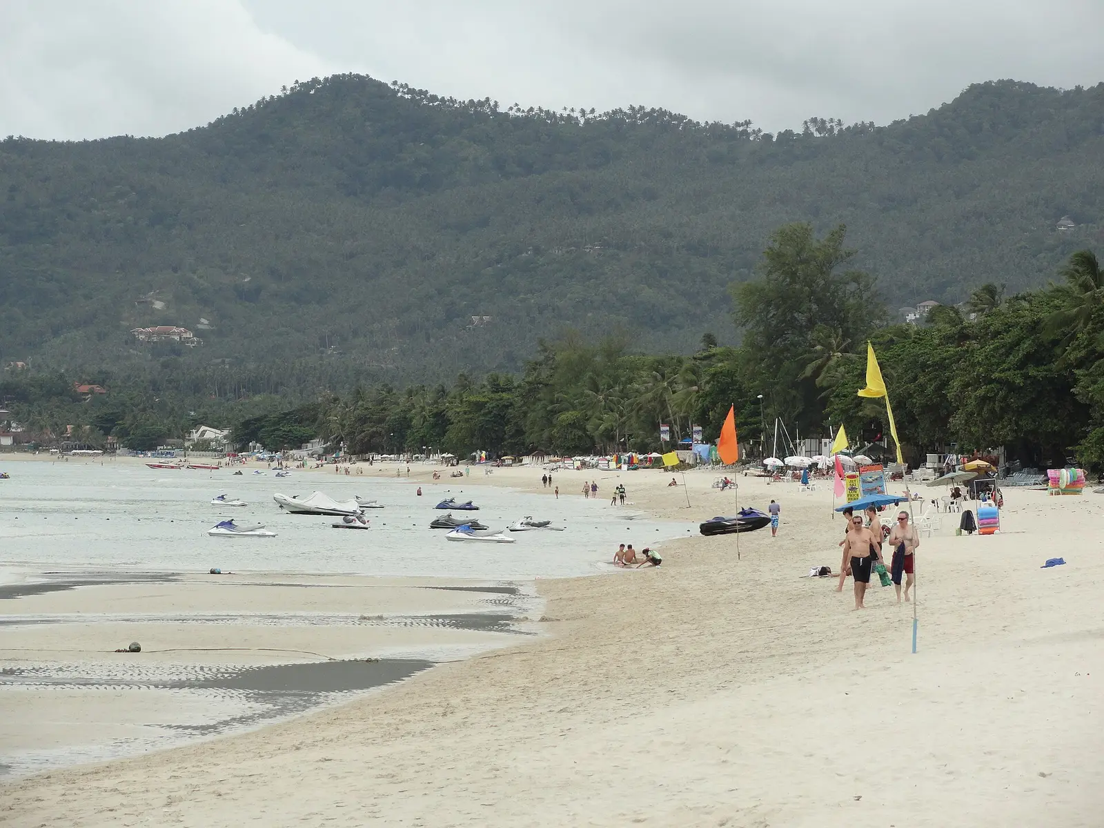 Sailing boats and beachgoers on the sand of Chaweng Beach with green hills behind
