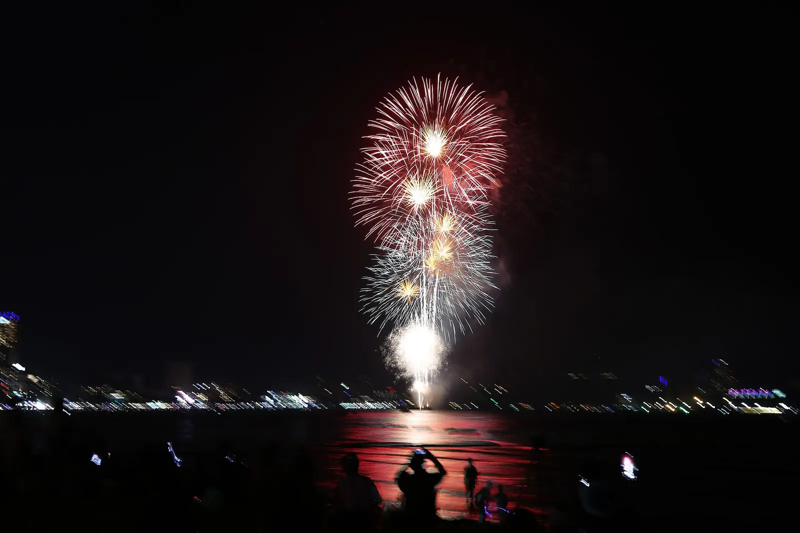 Red and white fireworks bursting over Pattaya Bay with the city lights along the shore